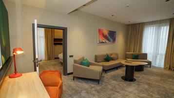 A hotel room with a sofa set, a coffee table, a desk with a lamp, and a painting on the wall.