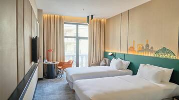 A hotel room with two beds, a desk with a chair, a lamp, and a view of a cityscape through the window.