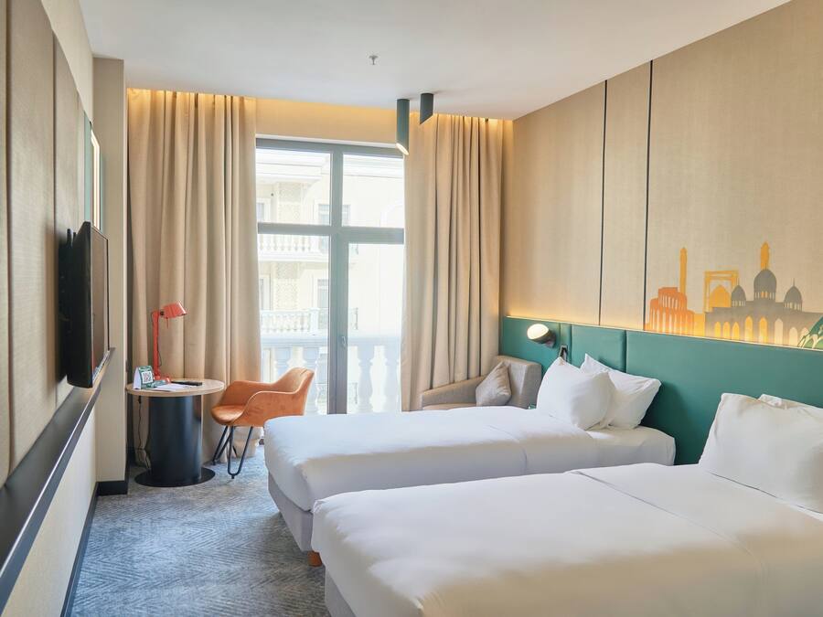 A hotel room with two beds, a desk with a chair, a lamp, and a view of a cityscape through the window.