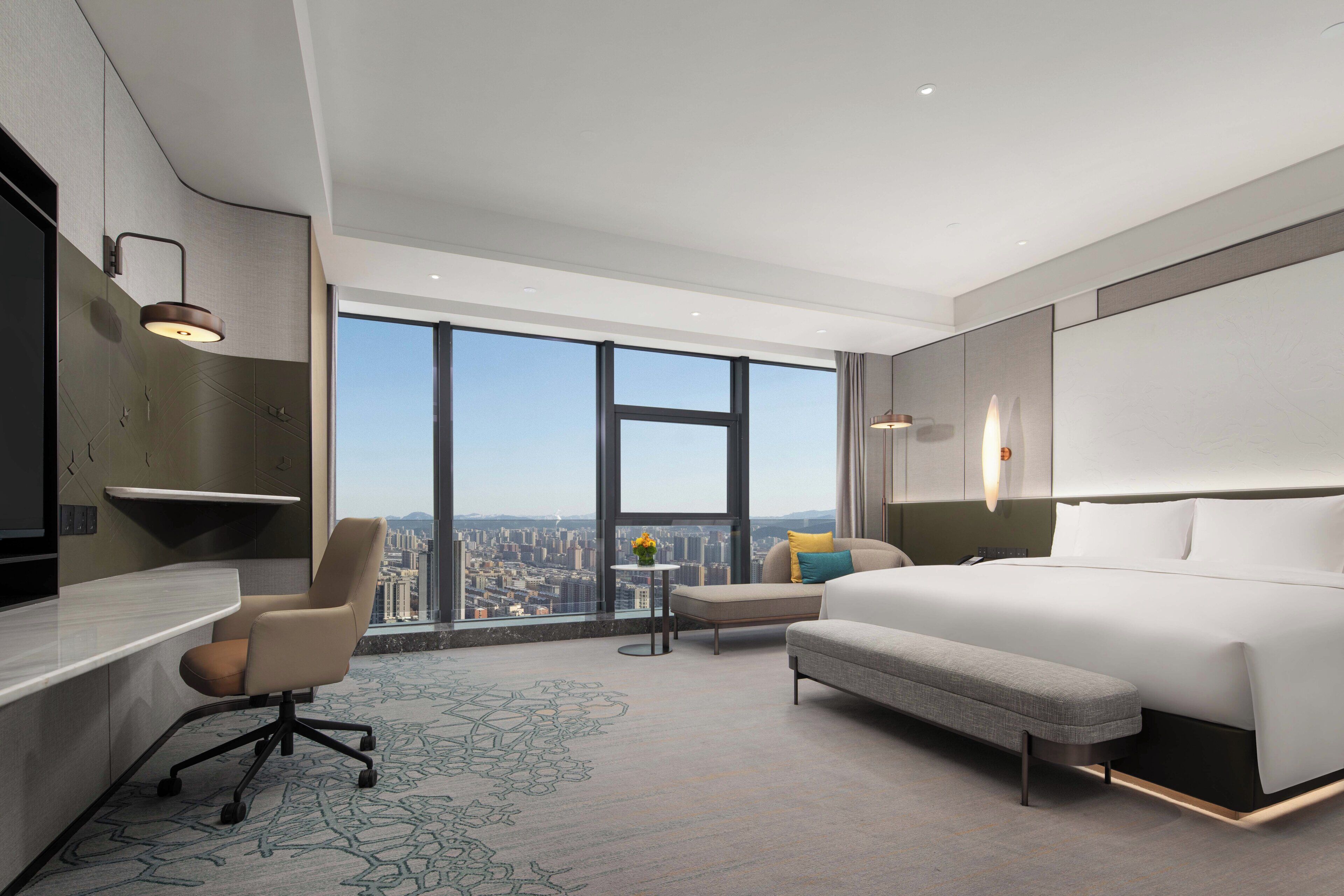 A modern hotel room with a large bed, a desk, a chair, and a view of the city.