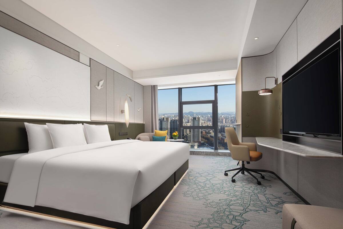 A modern hotel room with a large bed, a desk with a flat-screen TV, and a city view through a large window.