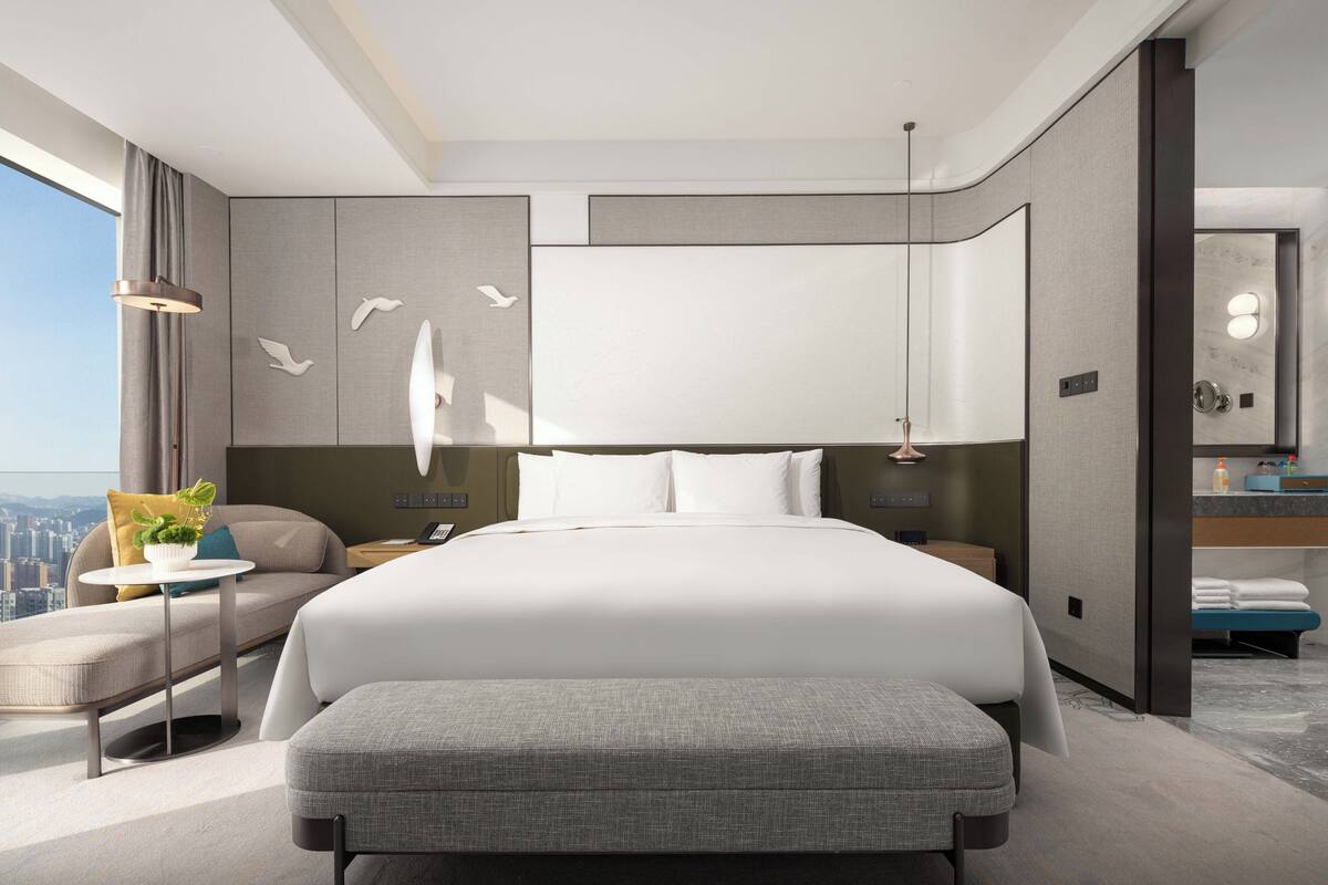 A modern hotel room with a large bed, a seating area, a small table, and a view of the city.