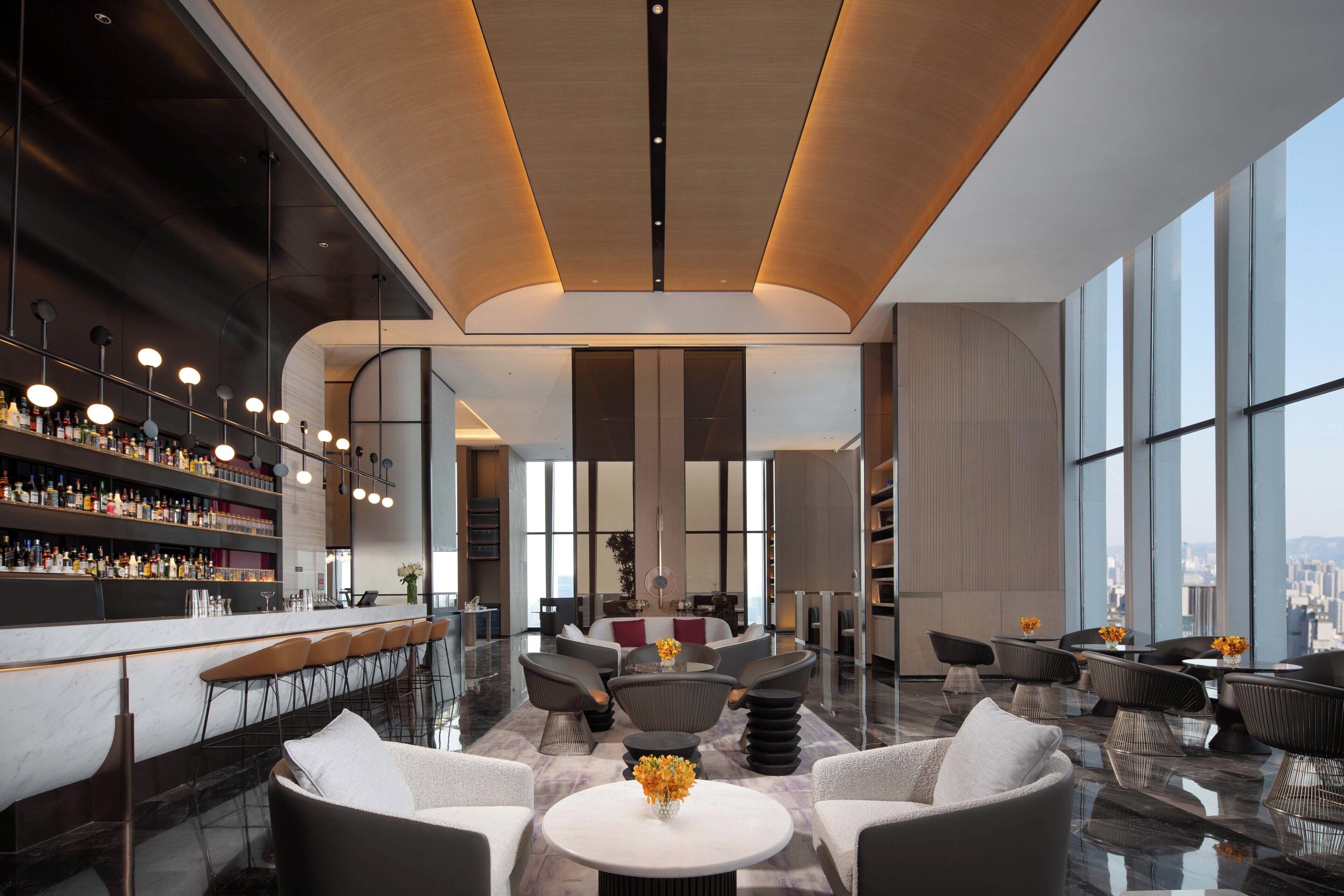 Un bar moderne doté d’un long comptoir, de tabourets et d’une grande variété de bouteilles. On y trouve également des espaces salon avec des tables et des chaises rondes, ainsi que de grandes fenêtres offrant une vue sur la ville.