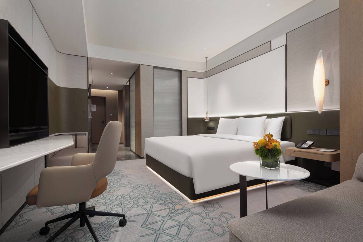 A modern hotel room with a large bed, a desk, a chair, a small table with flowers, and a bedside lamp.