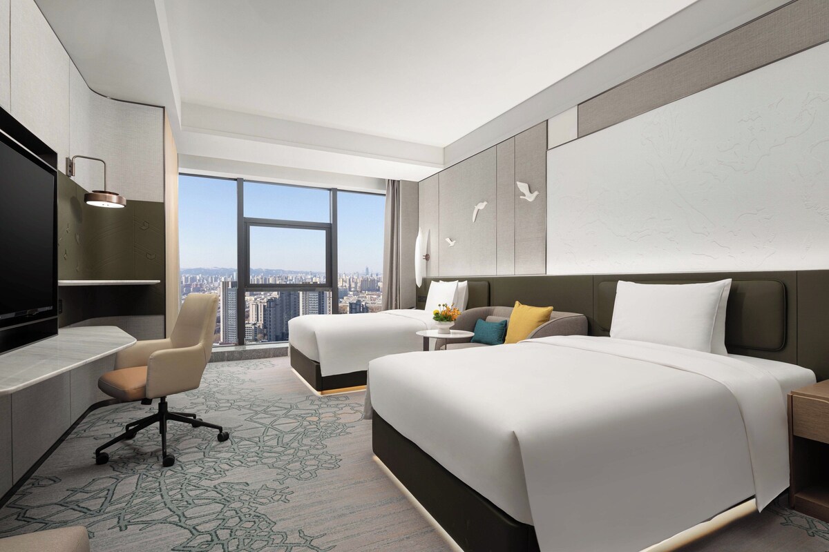 Habitación de hotel con dos camas, un escritorio, una silla y un amplio ventanal con vistas a la ciudad.