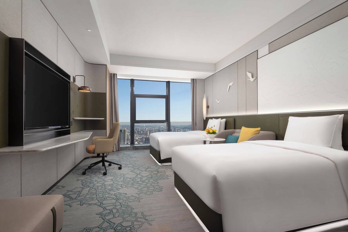 A hotel room with two beds, a TV, a desk, and a view of the city.