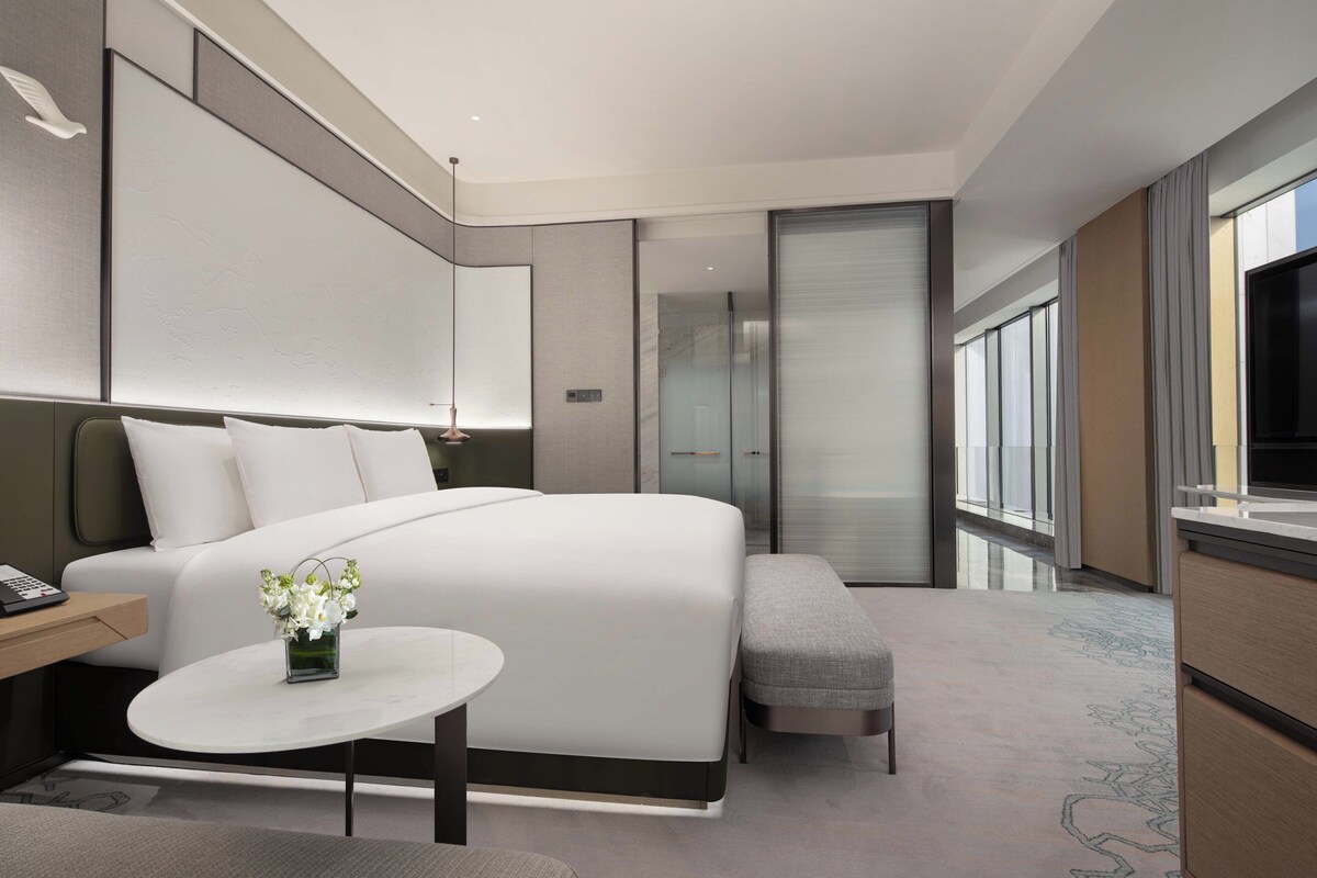 Habitación de hotel moderna con una cama grande, una mesita con un jarrón de flores, un banco y un amplio ventanal con vistas.