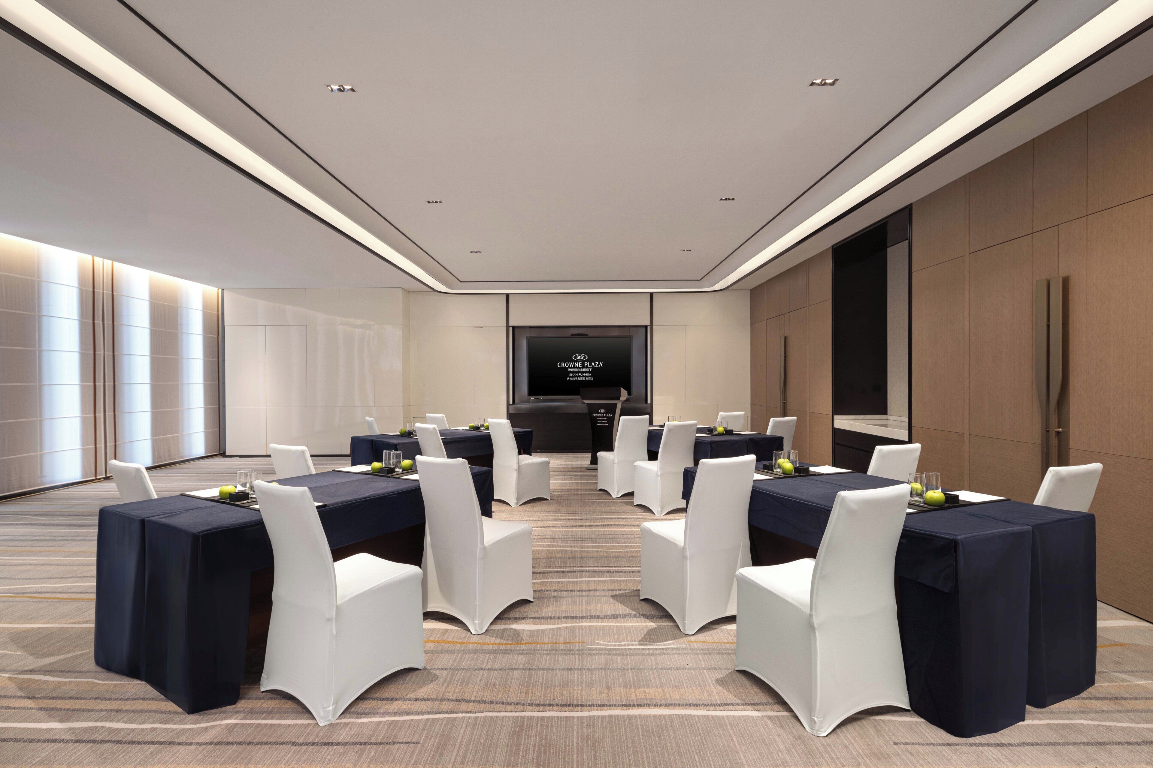 Un salón de conferencias con una larga mesa, sillas blancas y una pantalla al frente.