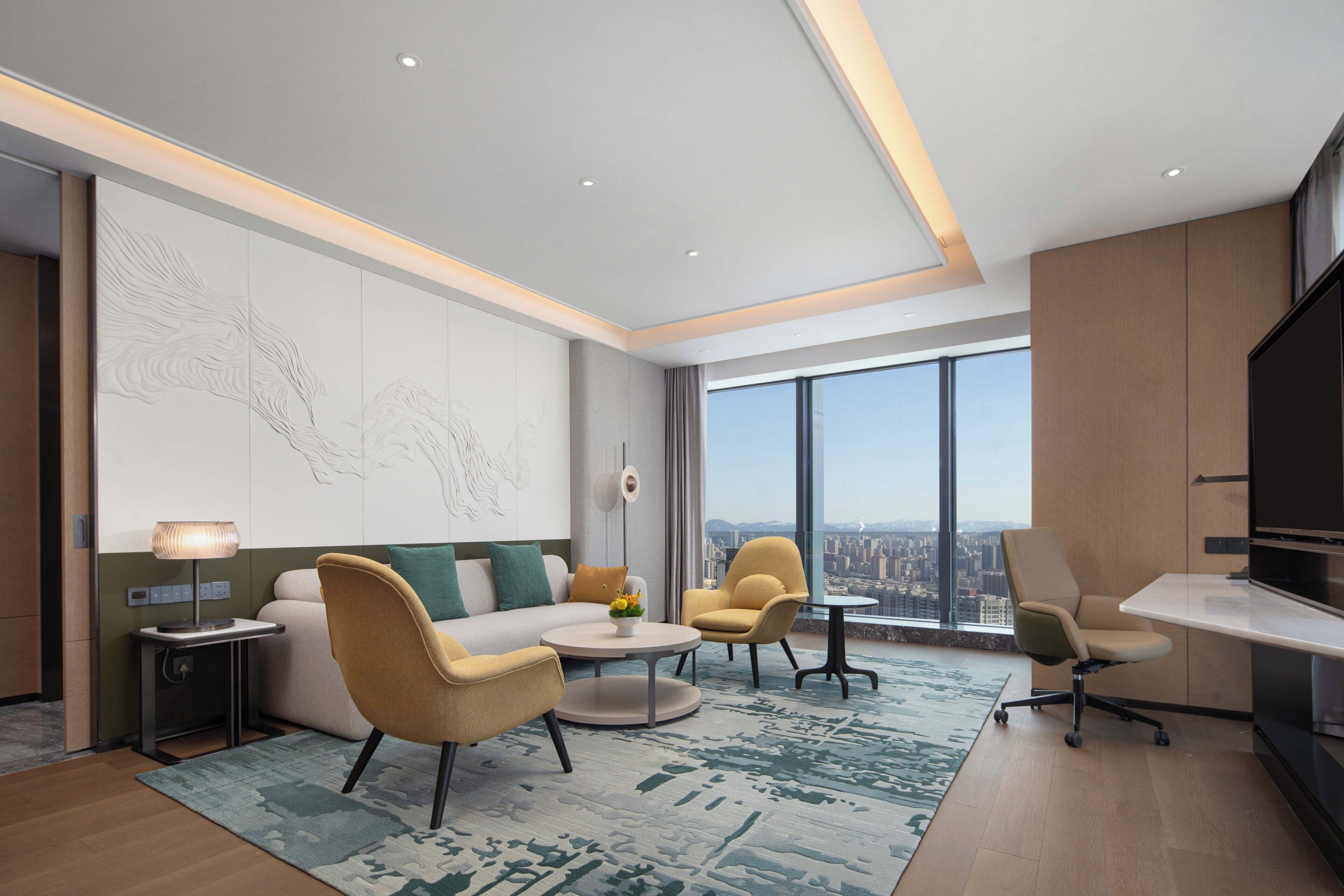 A modern living room with a sofa, two armchairs, a coffee table, and a desk. There is a large window with a city view, a decorative wall piece, and a flat-screen TV.