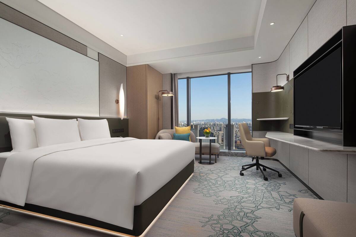 A modern hotel room with a large bed, a desk, a chair, a TV, and a view of the city.