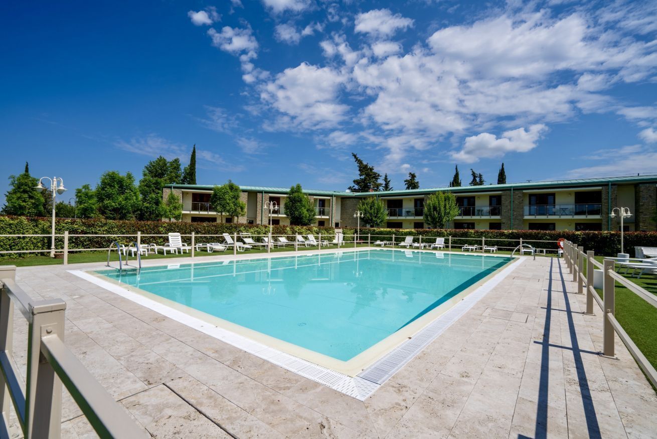 A rectangular pool with a tiled deck, surrounded by lounge chairs and a building with balconies in the background.