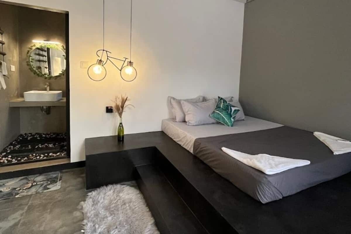 Een moderne slaapkamer met een bed, een klein zithoekje, een kaptafel met spiegel, en een bijzondere lamp in de vorm van een fiets.
