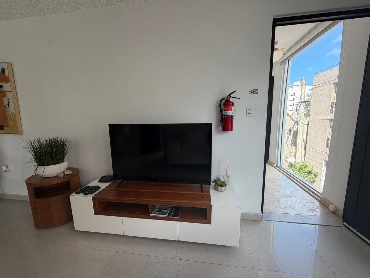 Een moderne woonkamer met een flatscreen televisie op een houten console, een kleine plant en een brandblusser aan de muur.