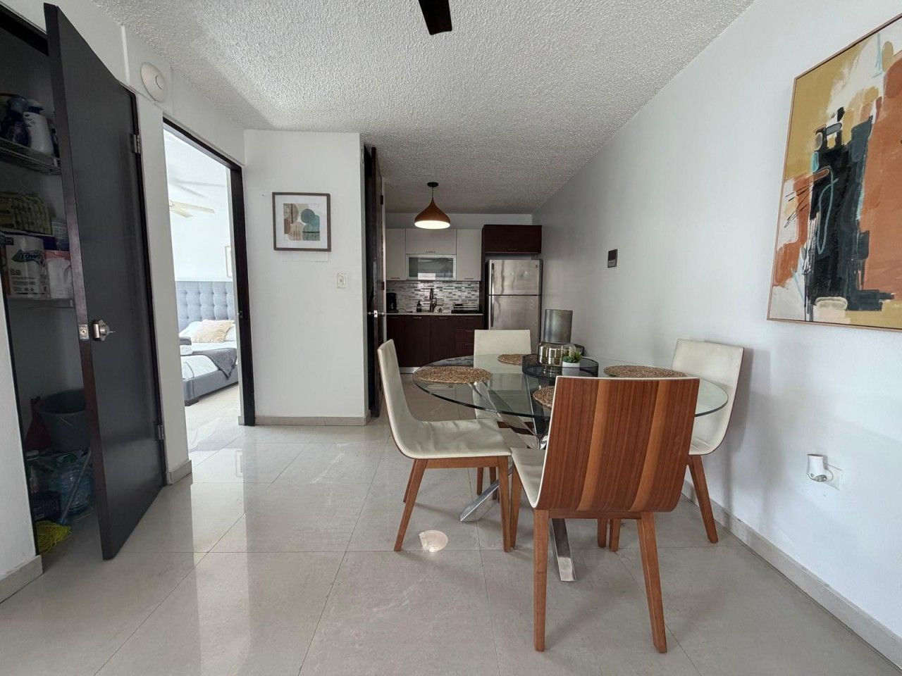 Un comedor moderno con una mesa y sillas de vidrio, una cocina al fondo y un cuadro en la pared.