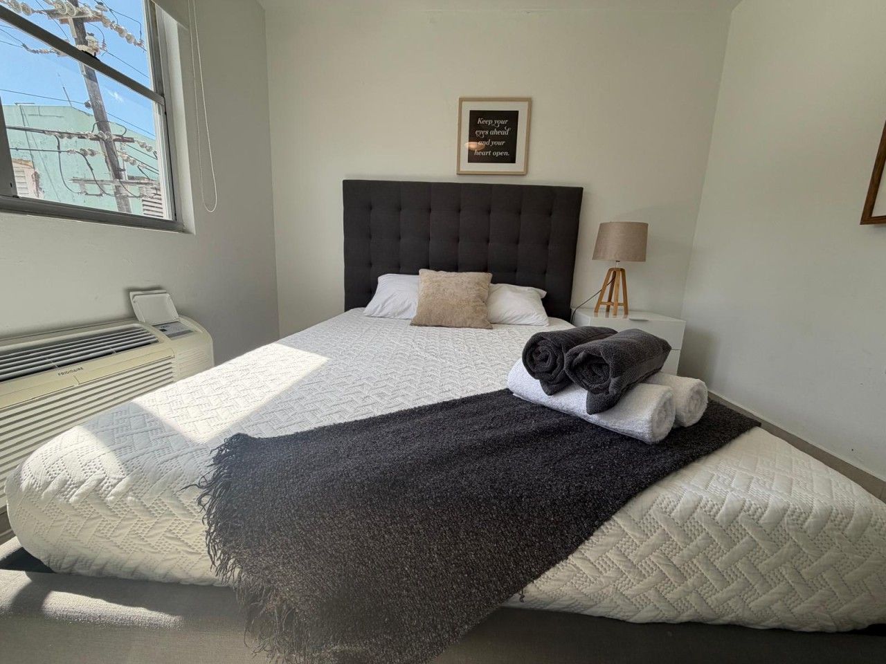 Een slaapkamer met een bed, een bedlampje, een citaat in een lijst op de muur en een raam met uitzicht op elektriciteitskabels.
