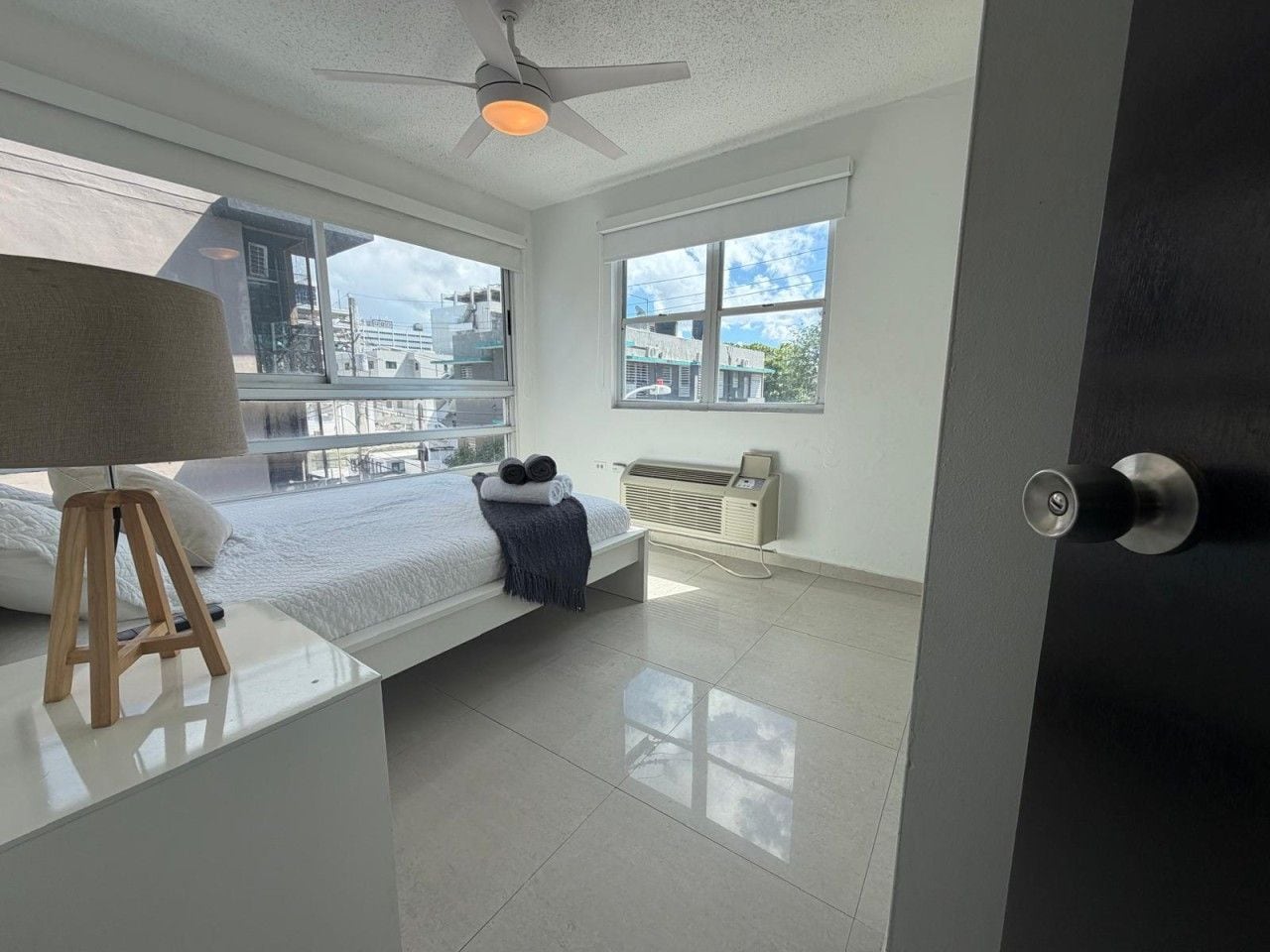 Quarto moderno com cama, mesa de cabeceira com abajur, radiador e janela com vista para prédios.