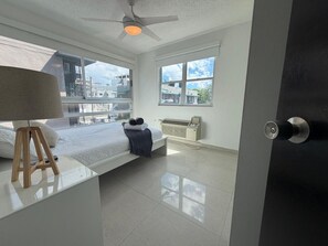 A modern bedroom with a bed, a bedside table with a lamp, a radiator, and a window with a view of buildings. - Bright, Comfy Apt @ Miramar (San Juan)