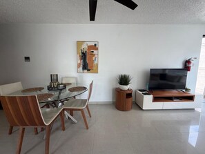 A modern living room with a glass dining table, wooden chairs, a flat-screen TV, and a painting on the wall.
