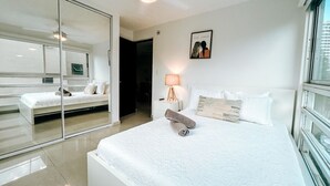 A modern hotel room with a large bed, a mirror, a bedside lamp, and a view of the city through the window. - Bright, Comfy Apt @ Miramar (San Juan)