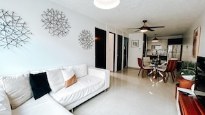 A modern living room with a white sofa, a dining area with chairs, a ceiling fan, and a kitchen in the background. - Bright, Comfy Apt @ Miramar (San Juan)