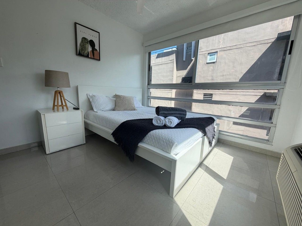 Une chambre à coucher avec un lit, une table de chevet, une lampe, une fenêtre et un radiateur.