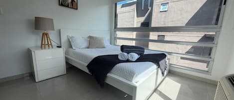 Une chambre à coucher avec un lit, une table de chevet, une lampe, une fenêtre et un radiateur.