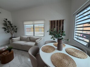 Una sala de estar moderna con un sofá, una mesa de comedor con manteles individuales, una planta en maceta y vistas amplias por las ventanas.