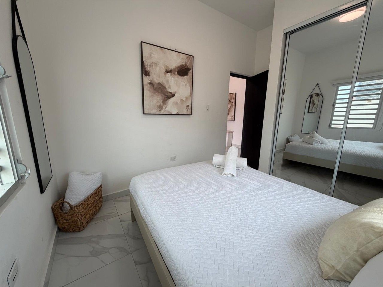 A bedroom with a bed, a wall-mounted abstract painting, a wicker basket, and a large mirror reflecting a bed and a window.
