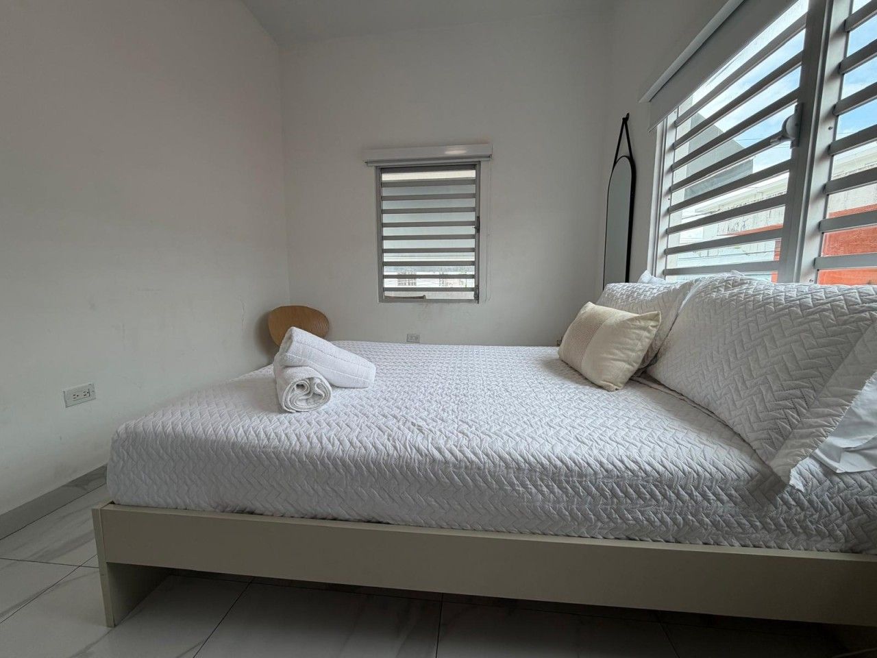 Ein Zimmer mit einem Einzelbett, einer weißen Bettdecke, einem kleinen Fenster mit Jalousien und einem Nachttisch mit Lampe.