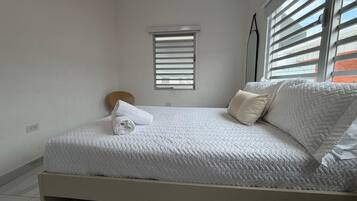 Ein Zimmer mit einem Einzelbett, einer weißen Bettdecke, einem kleinen Fenster mit Jalousien und einem Nachttisch mit Lampe.