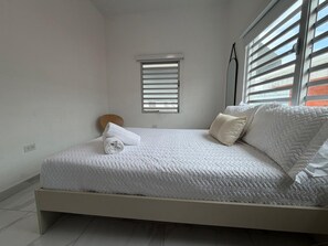 Habitación con una cama individual, cubrecama blanco, un ventanal pequeño con persianas y una mesita de noche con lámpara.