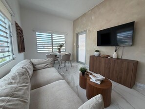 Un salon moderne avec un canapé d’angle, un coin repas avec des chaises, un téléviseur à écran plat fixé au mur et un meuble en bois.
