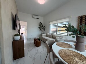 A modern living room with a sofa, a dining table with placemats, a TV mounted on the wall, and a potted plant.