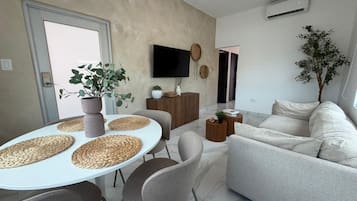 Ein modernes Wohnzimmer mit einer Couch, einem Esstisch mit Unterlagen, einem Fernseher und einer Topfpflanze.