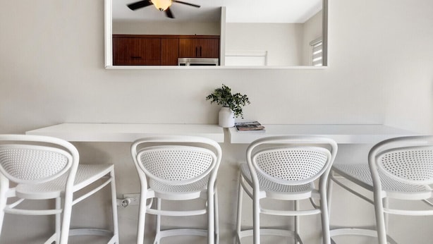 Un coin repas avec une table blanche et quatre chaises blanches, un ventilateur de plafond et une petite plante en pot posée sur la table.