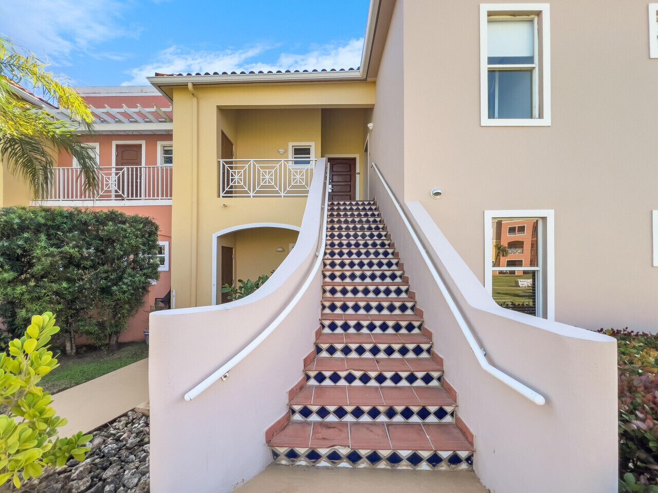 A staircase leading to a building entrance with a railing, surrounded by greenery and a clear sky.