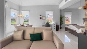 A modern living room with a beige sofa, a dining area with a table and chairs, a workspace with a desk and chair, and a bedroom with a bed and a nightstand. - Cozy Beach Front Apt @ Palmas del Mar w/ 2 Pools (Palmas del Mar)