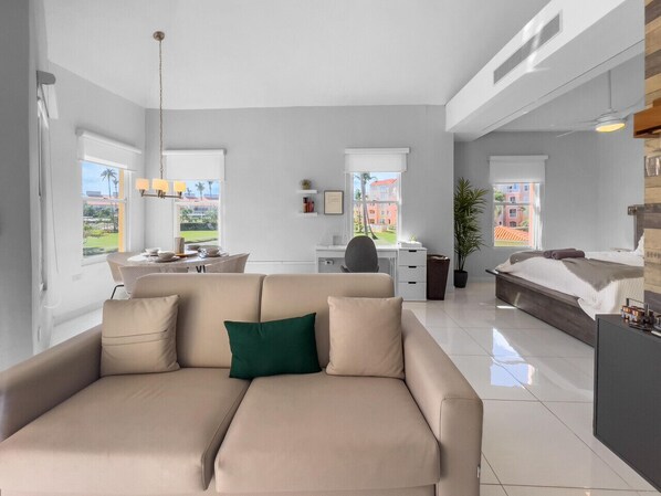 A modern living room with a beige sofa, a dining area with a table and chairs, a workspace with a desk and chair, and a bedroom with a bed and a nightstand.
