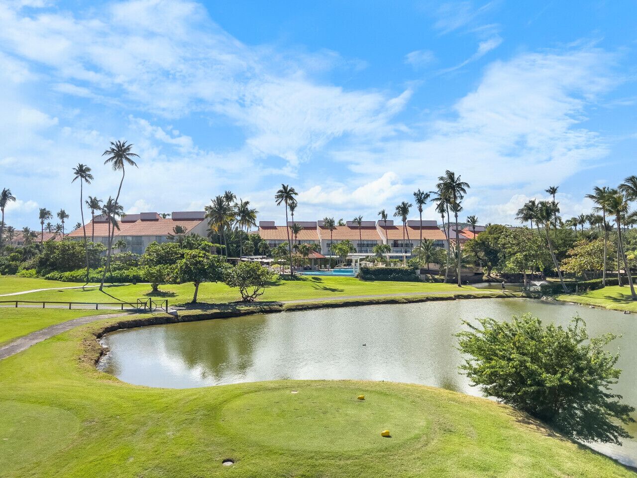 Ein Golfplatz mit Teich, Palmen und einem Gebäude mit rotem Dach im Hintergrund.