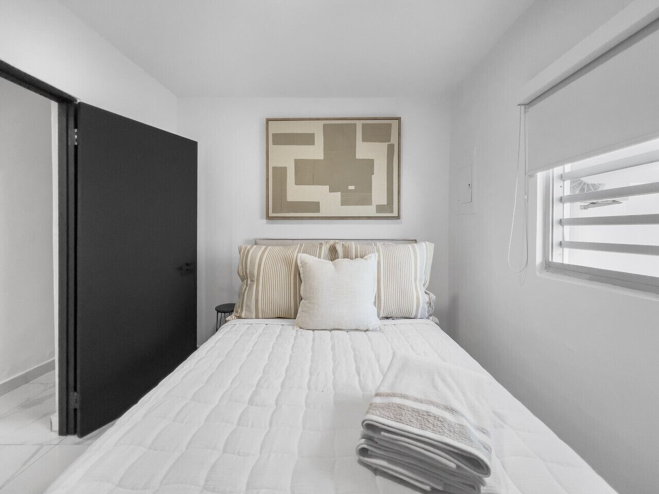 A bedroom with a bed, pillows, a framed abstract artwork, a window with blinds, and a door.