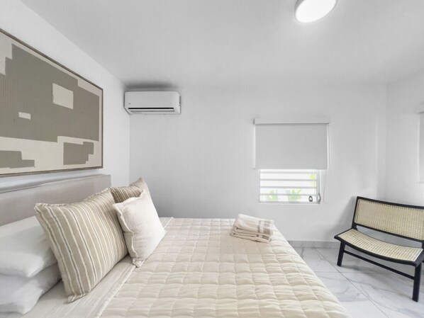 A bedroom with a bed, pillows, a bench, a wall-mounted air conditioner, and a window with blinds.