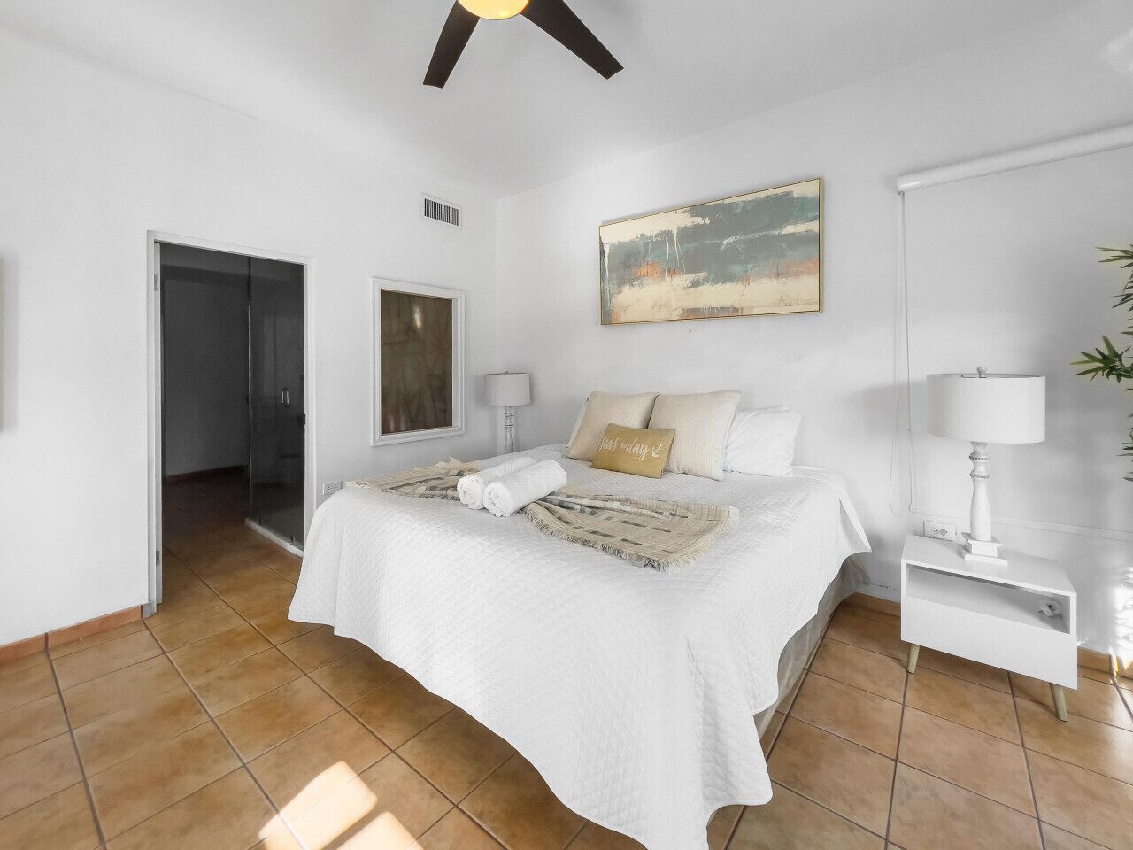 Un dormitorio con cama, mesita de noche, ventilador de techo, un cuadro y una puerta que da a otra habitación.