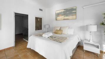 Un dormitorio con cama, mesita de noche, ventilador de techo, un cuadro y una puerta que da a otra habitación.