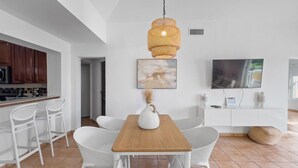 A modern kitchen with white cabinetry, a wooden dining table, and white chairs. There is a hanging wicker light fixture above the table and a flat-screen TV mounted on the wall. - Oasis Beach Apt in Palmas del Mar w/ Pool & Views (Palmas del Mar)