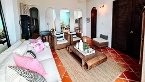 Ein geräumiges Wohnzimmer mit einem weißen Sofa, Korbstühlen, einem Holztisch und einem roten Fliesenboden.