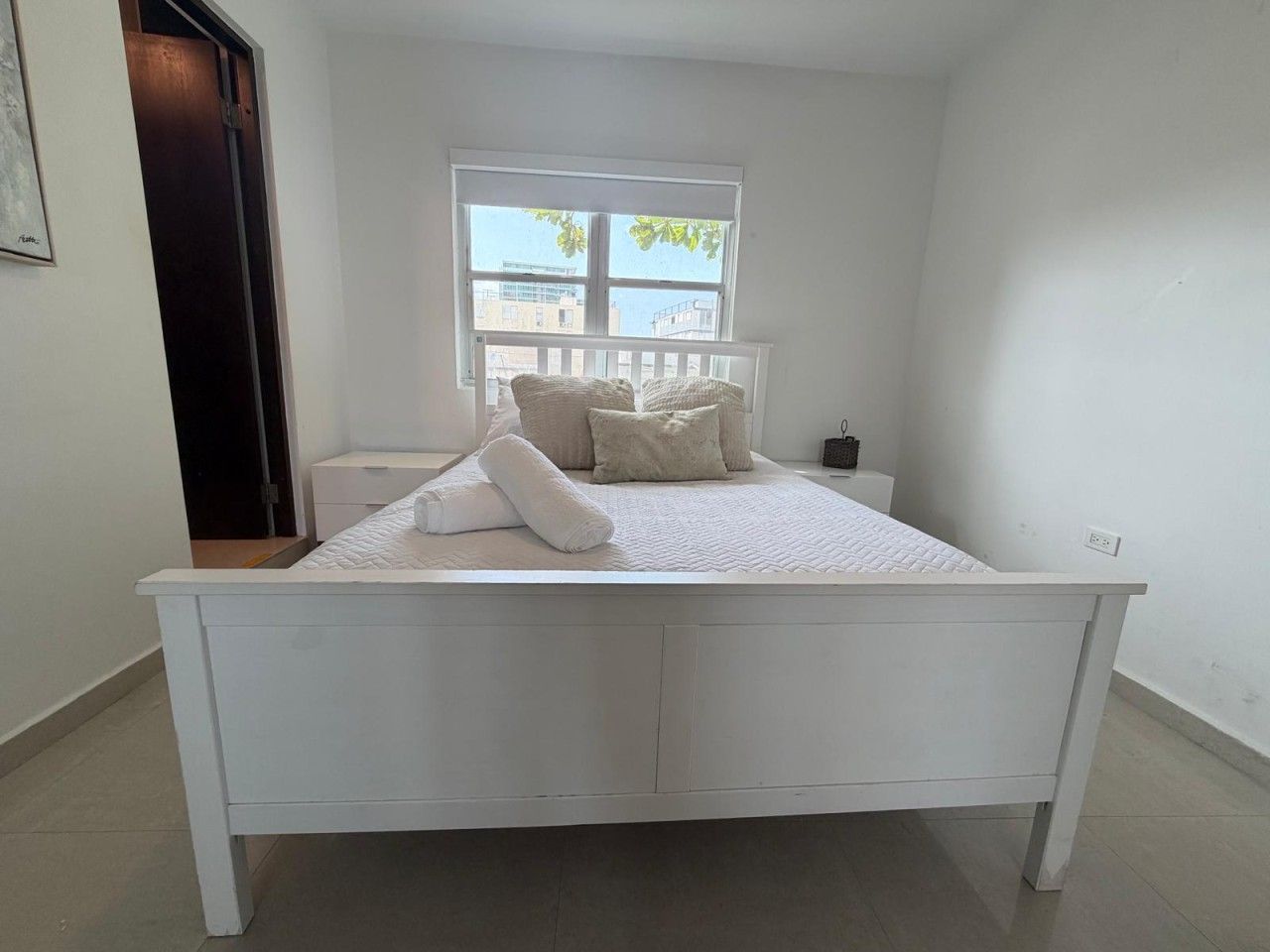 A white bed in a room with a window, a nightstand, and a door.