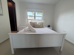 Una cama blanca en una habitación con ventana, una mesita de noche y una puerta.