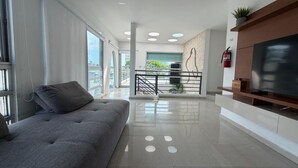 A modern living room with a grey sofa, a flat-screen TV on a wooden stand, and a balcony with a view. - Elegant Modern PH W/ Balcony & Parking (San Juan)