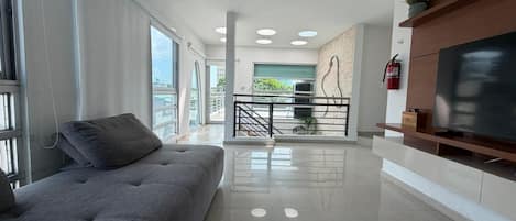 Een moderne woonkamer met een grijze bank, een flatscreen televisie op een houten standaard, en een balkon met uitzicht.