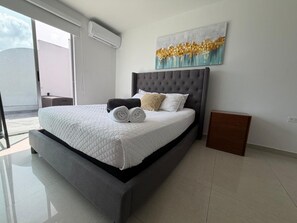 Une chambre moderne avec un grand lit, une tête de lit, une table de chevet, une commode, un climatiseur mural et une fenêtre donnant sur un beau paysage.